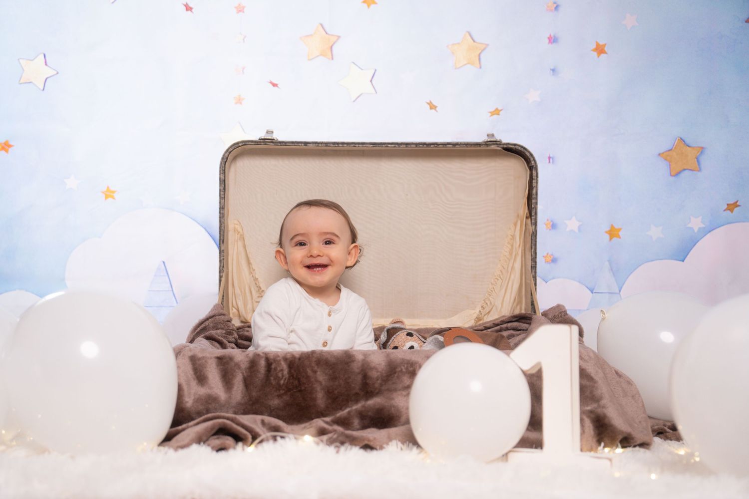 Baby sits in vintage suitcase surrounded by clouds and stars in dreamy first birthday photoshoot setup.