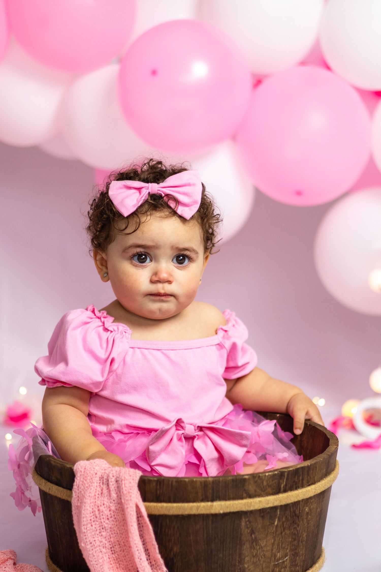 Baby sitting in wooden barrel wearing pink dress with bow against balloon backdrop.