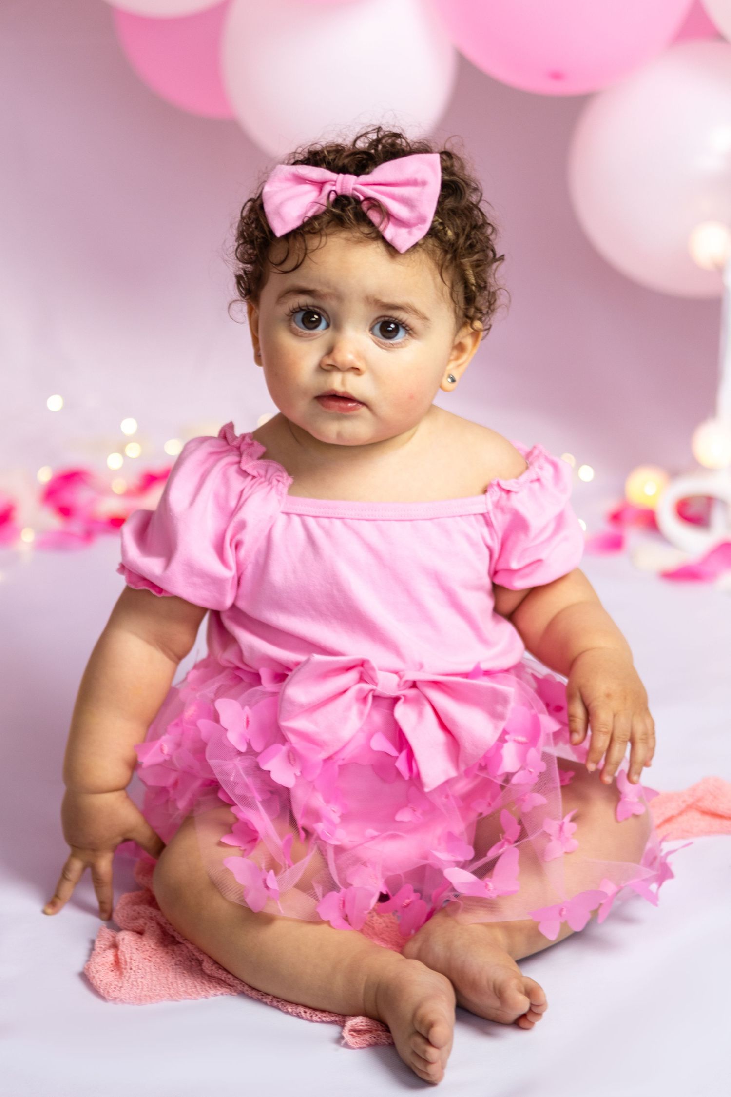 Baby in pink tutu dress sitting against pink balloon backdrop.