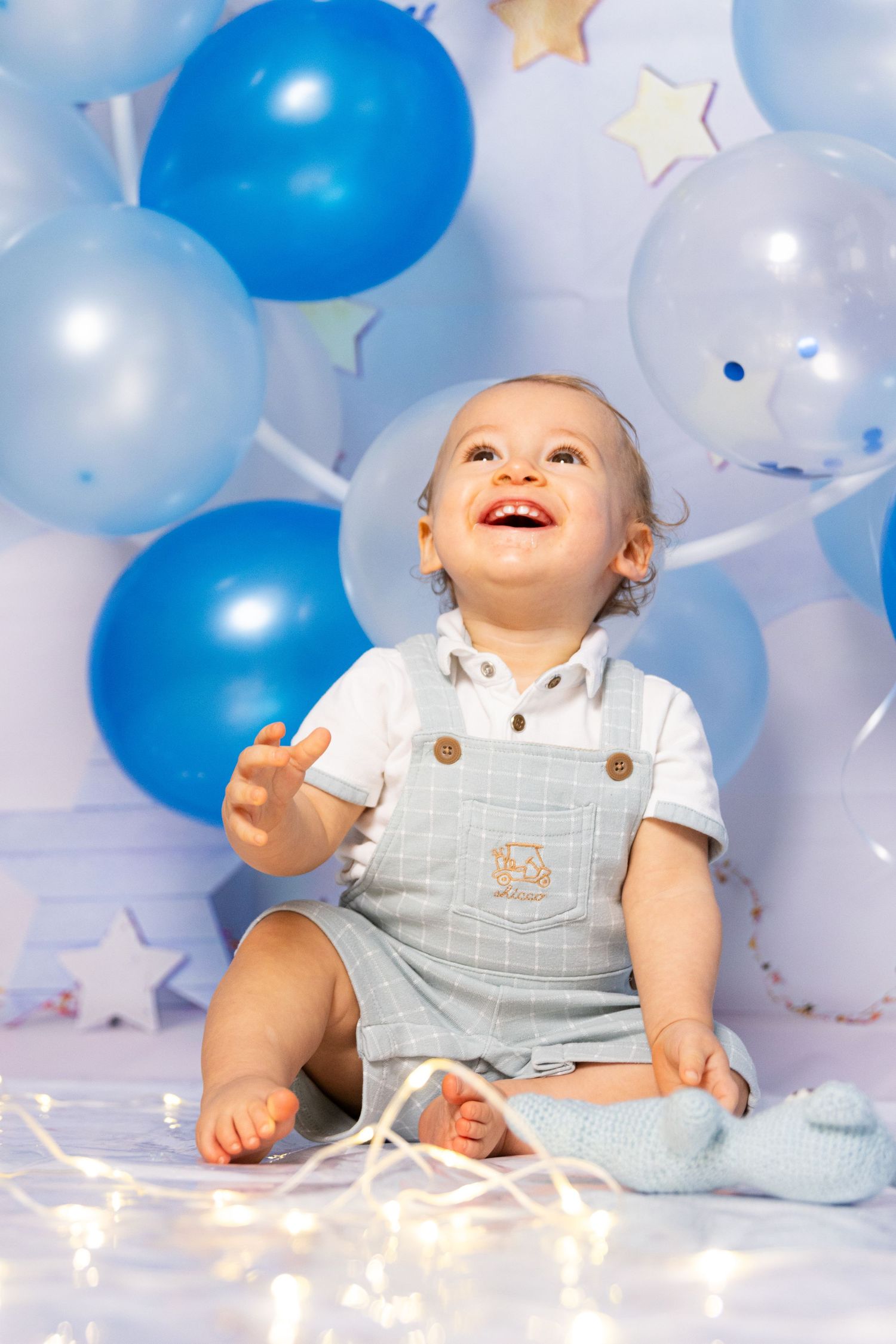Baby's first birthday photoshoot with blue and white balloons and string lights.