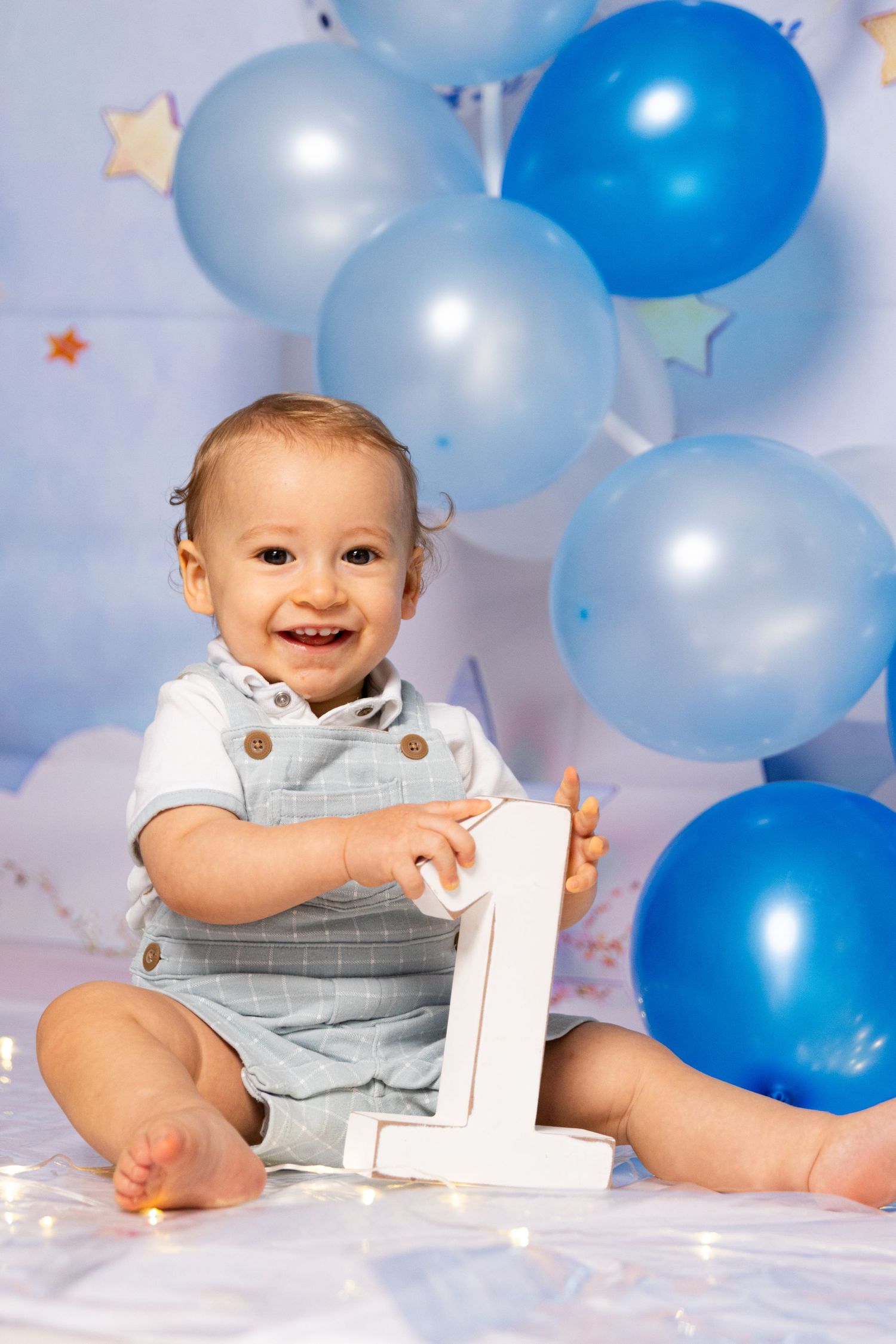 Smiling baby plays with number one prop amid blue balloons and twinkle lights.