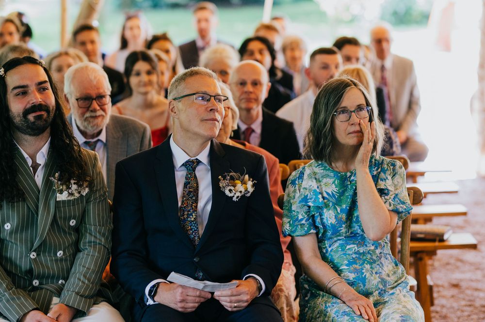 Wedding guests in formal attire sit in rows during a ceremony, with a woman wearing a bright blue floral dress.