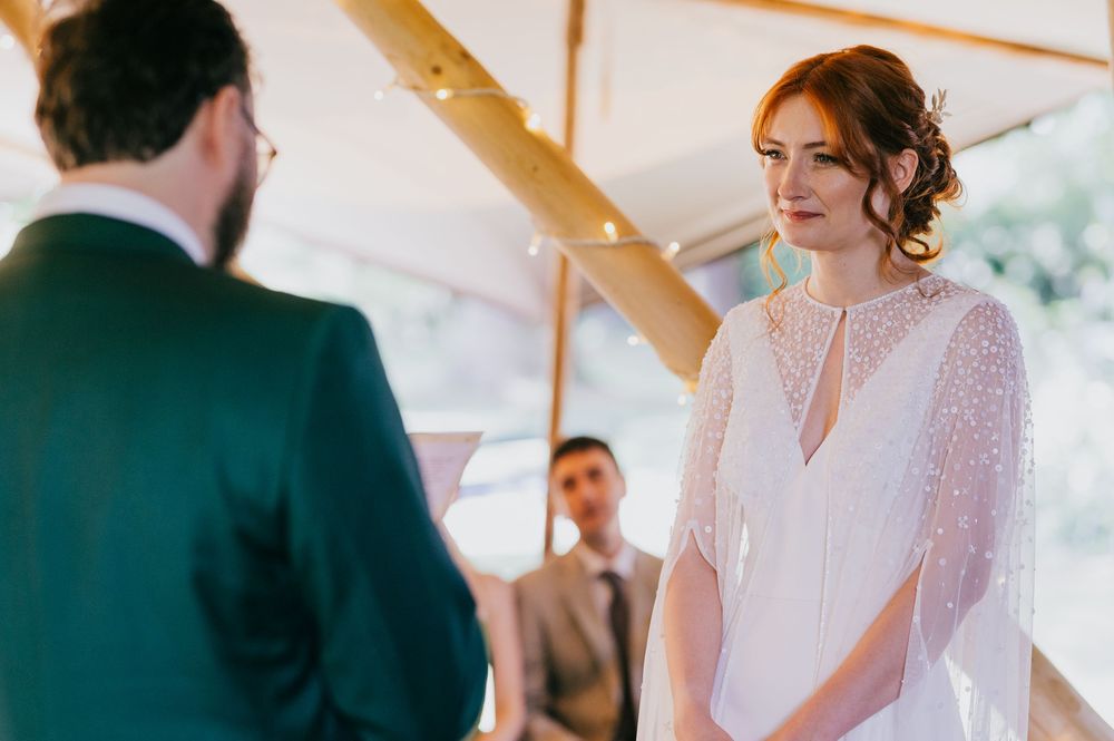 A bride in a delicate white lace dress faces the officiant during a wedding ceremony under a wooden structure.