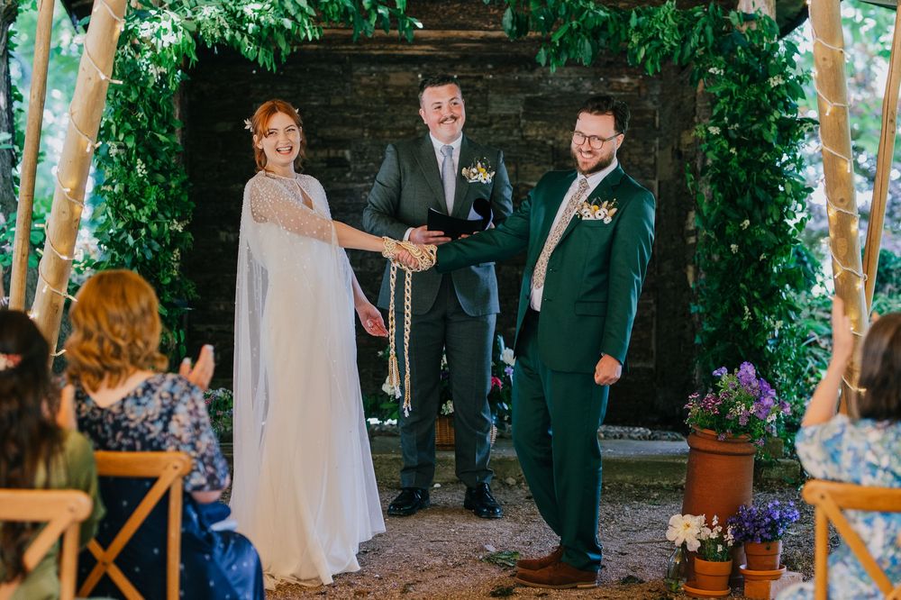 A romantic outdoor garden wedding ceremony with couple and officiant under a lush green archway with purple flowers.