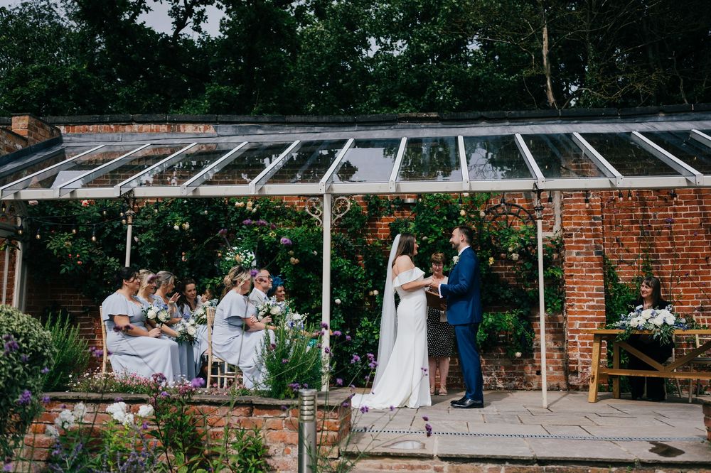 An outdoor wedding ceremony taking place under a glass conservatory roof against a rustic brick wall with guests seated nearby.