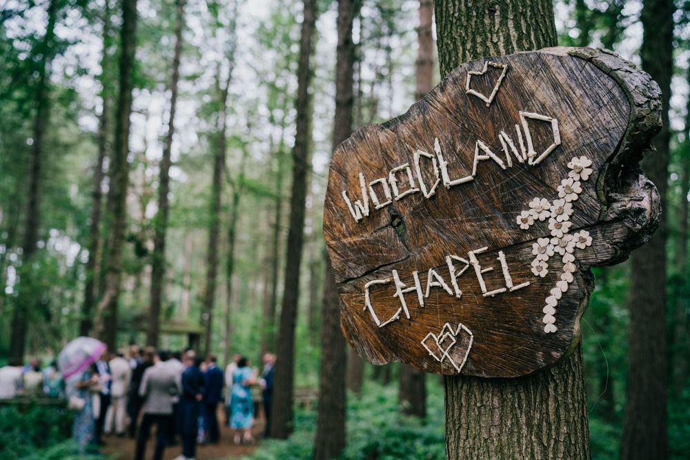Rustic wooden 'Woodland Chapel' sign mounted on tree trunk with crowd gathering in misty forest background.