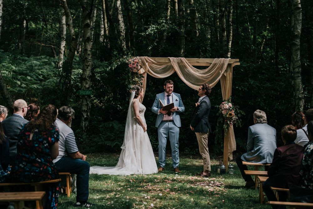 Guests seated at outdoor wooden benches during a rustic forest wedding ceremony under tall trees.