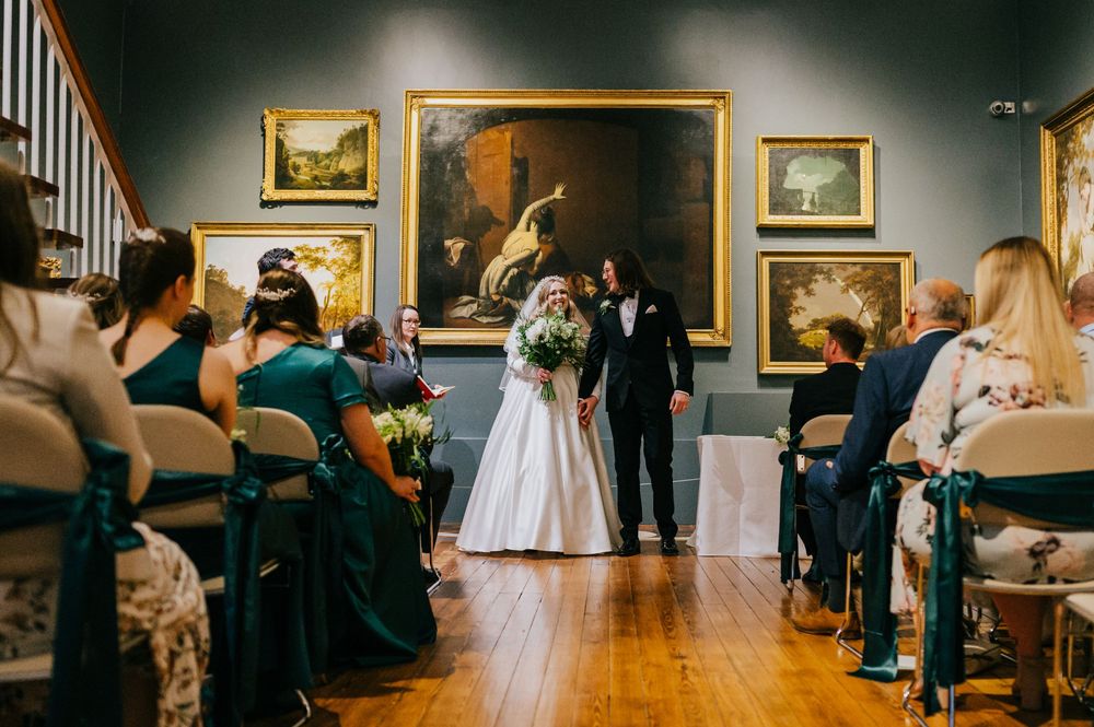 Wedding guests mingle in an elegant art gallery with golden-framed paintings adorning the walls during a reception.