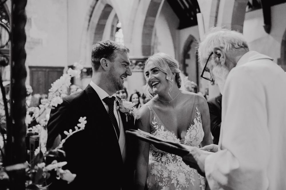 A black and white photograph captures a joyful candid moment during a wedding ceremony at a church.