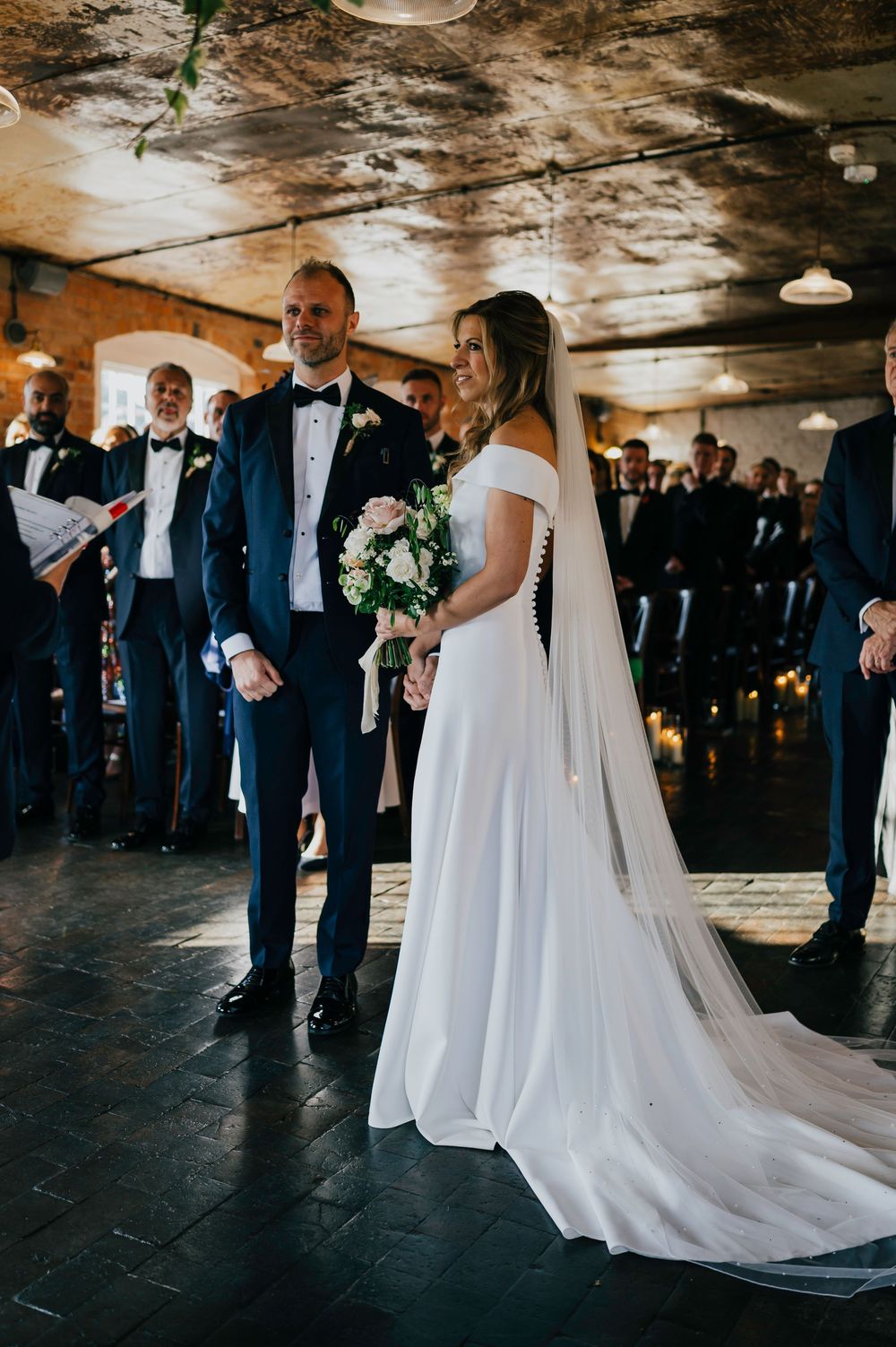 A bride in a white off-shoulder gown with long train stands next to a groom in navy suit during an indoor wedding ceremony.