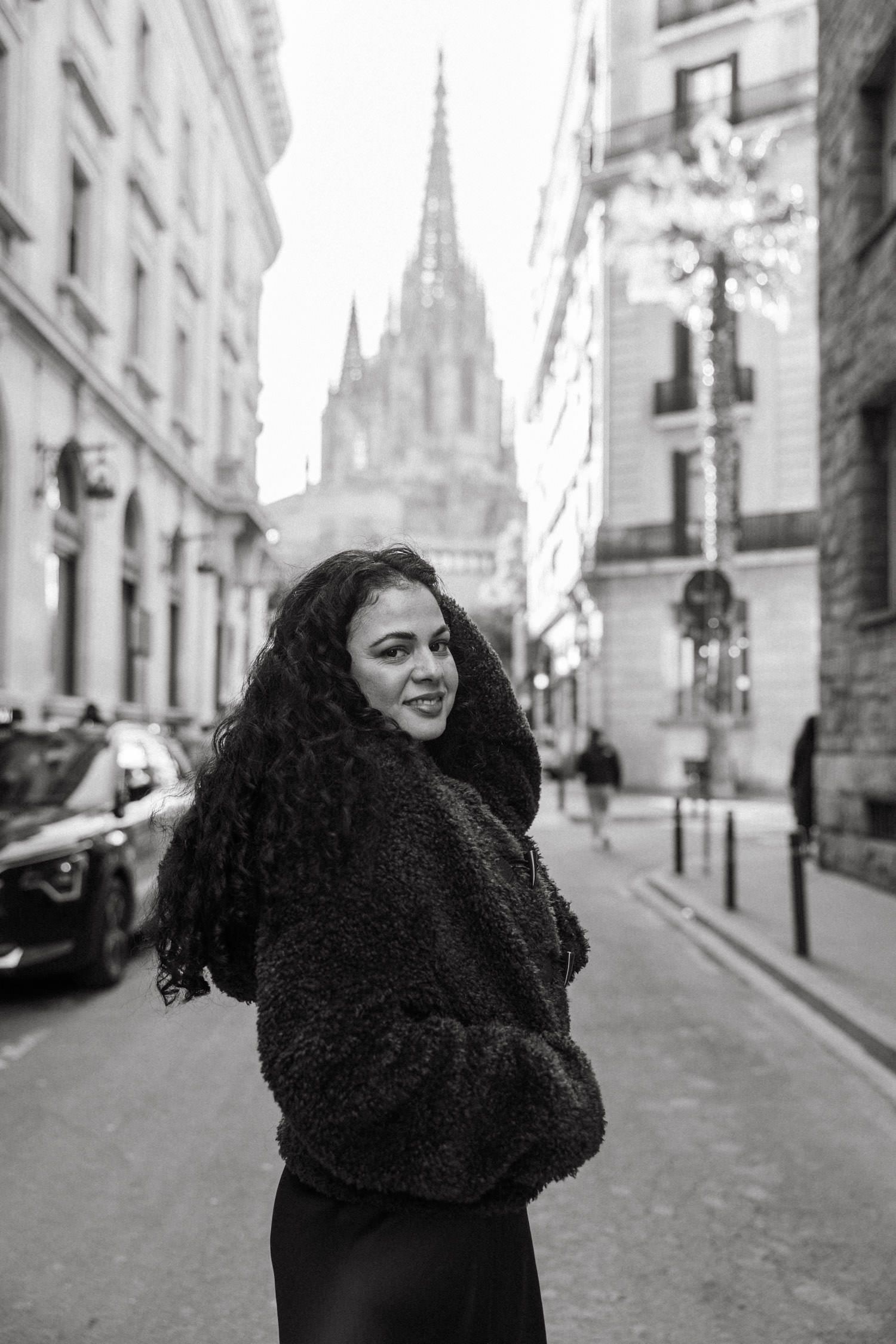 Woman in black coat poses playfully on cobblestone street with Gothic spires in background.