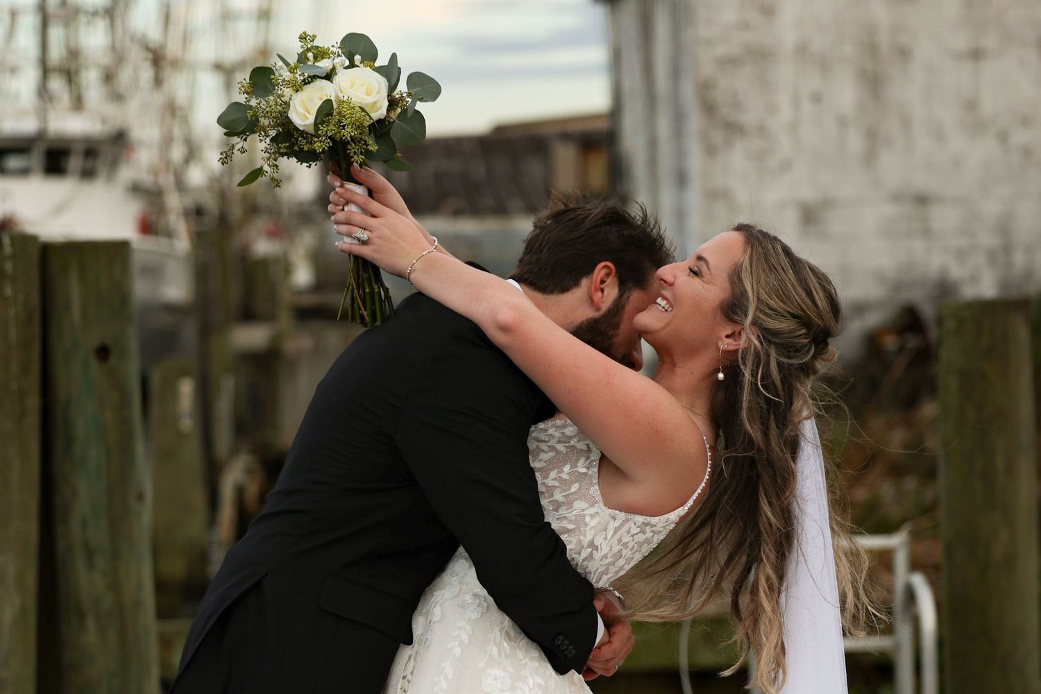 A wedding couple poses together on a wooden dock with fishing boats and cloudy skies in the background.