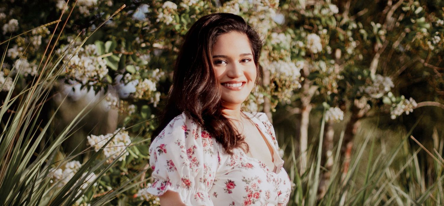 Smiling young woman in floral dress posing in front of white flowering bushes outdoors.