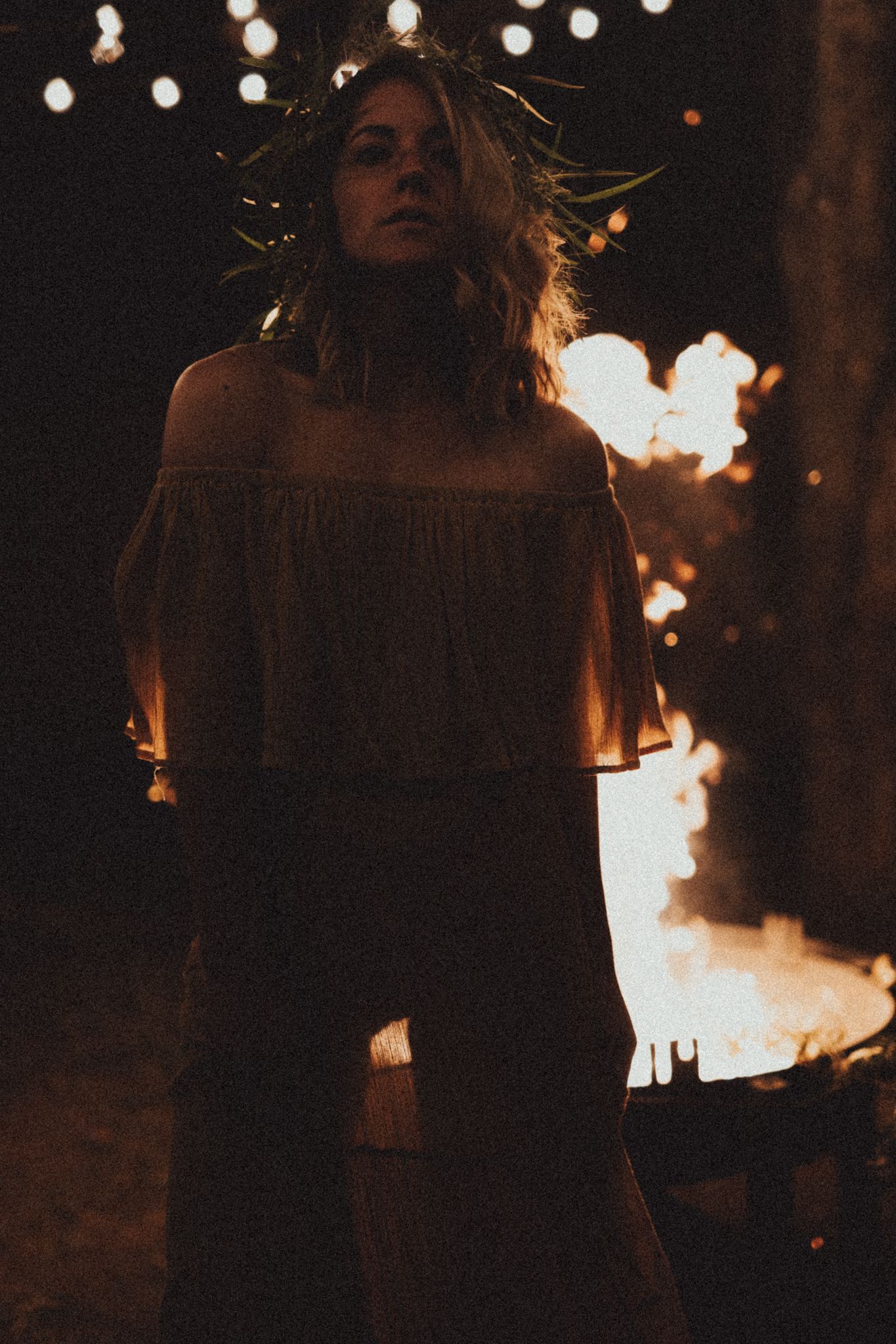 Person with long hair silhouetted against fairy lights and flames creating a moody nighttime atmosphere.