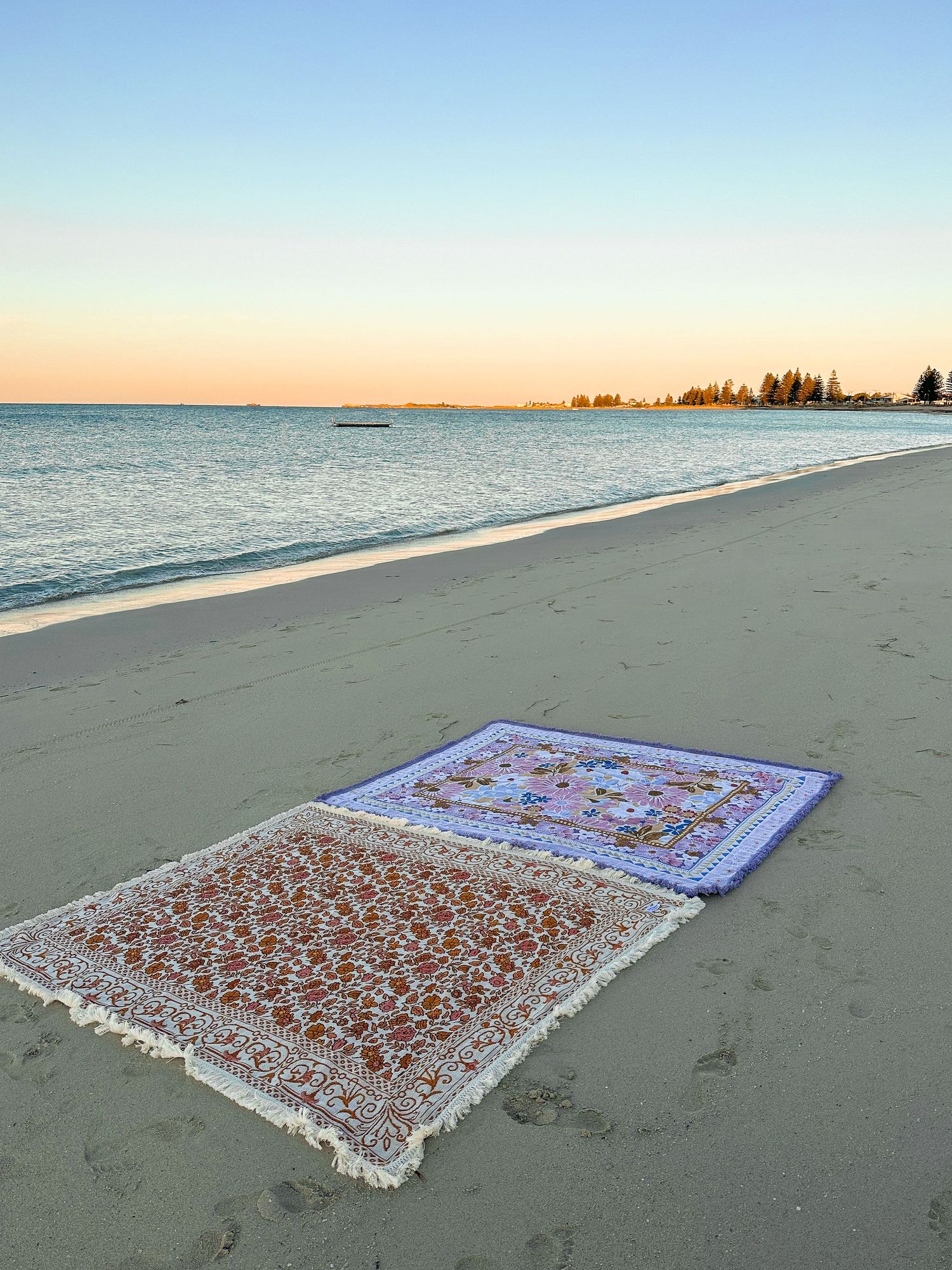 Two decorative beach blankets laid out on sand during sunset by the ocean.