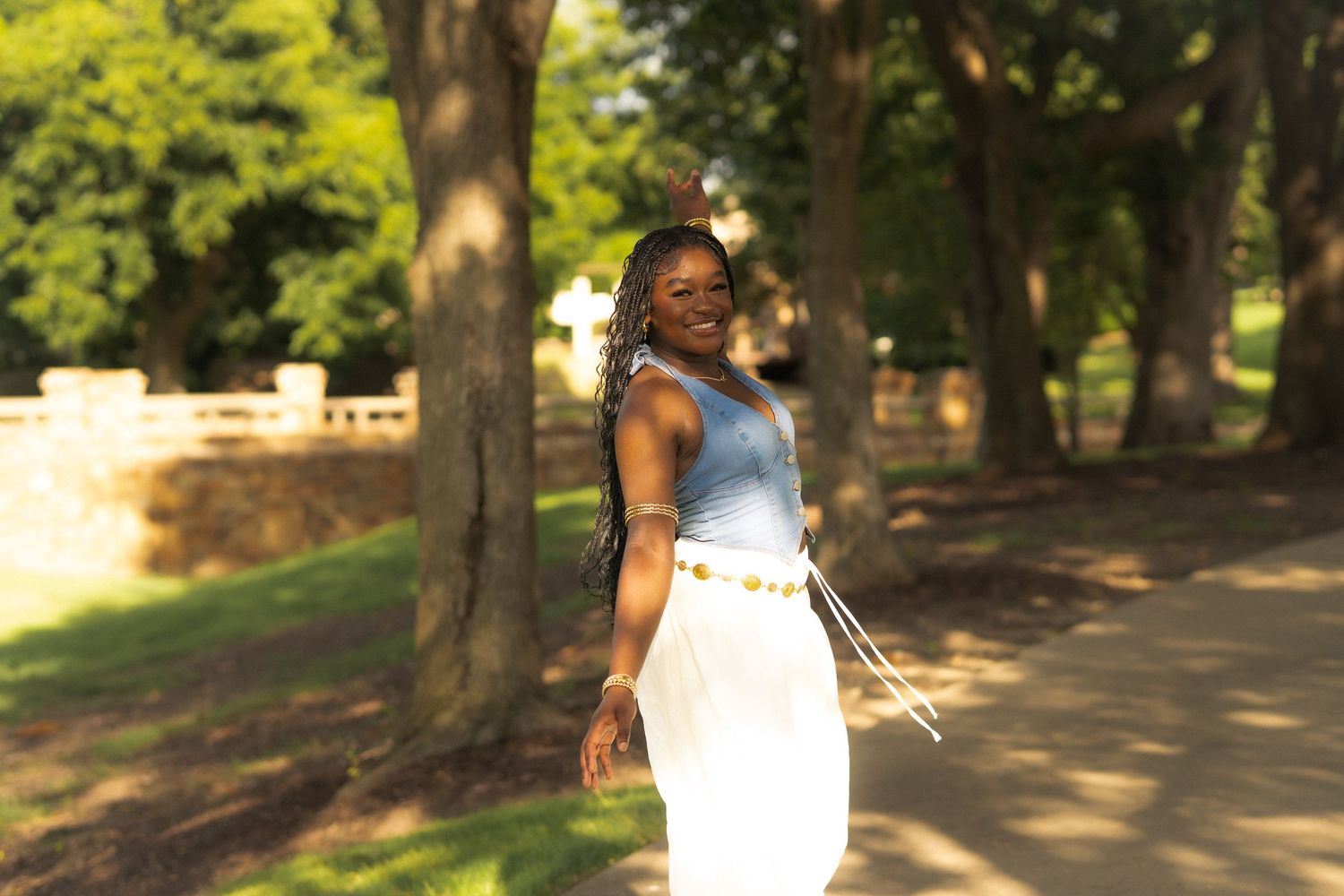 Someone in a white dress holds pink roses while smiling outdoors among green foliage on a sunny day.