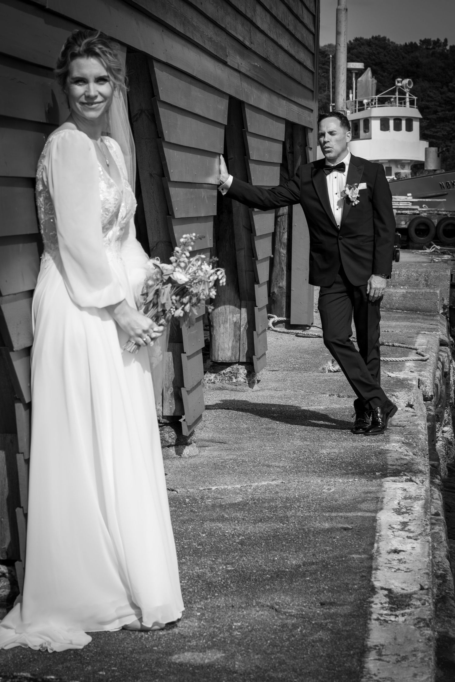 Bride in white gown holds bouquet while groom in tuxedo approaches along rustic waterfront building wall.