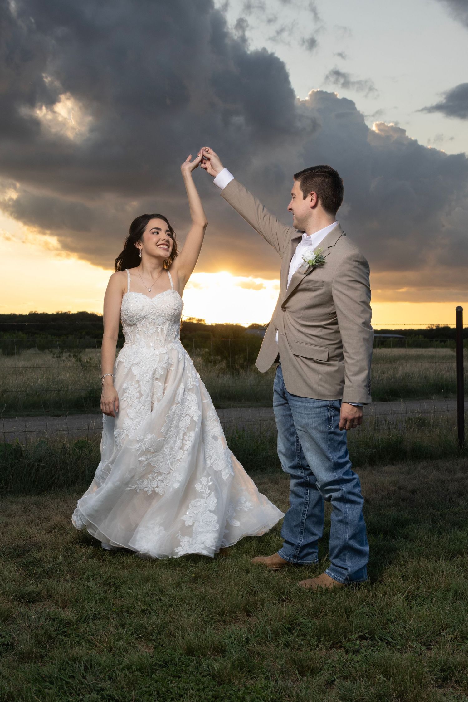 A couple dances together in a field during golden hour sunset, with the bride wearing a white lace gown.