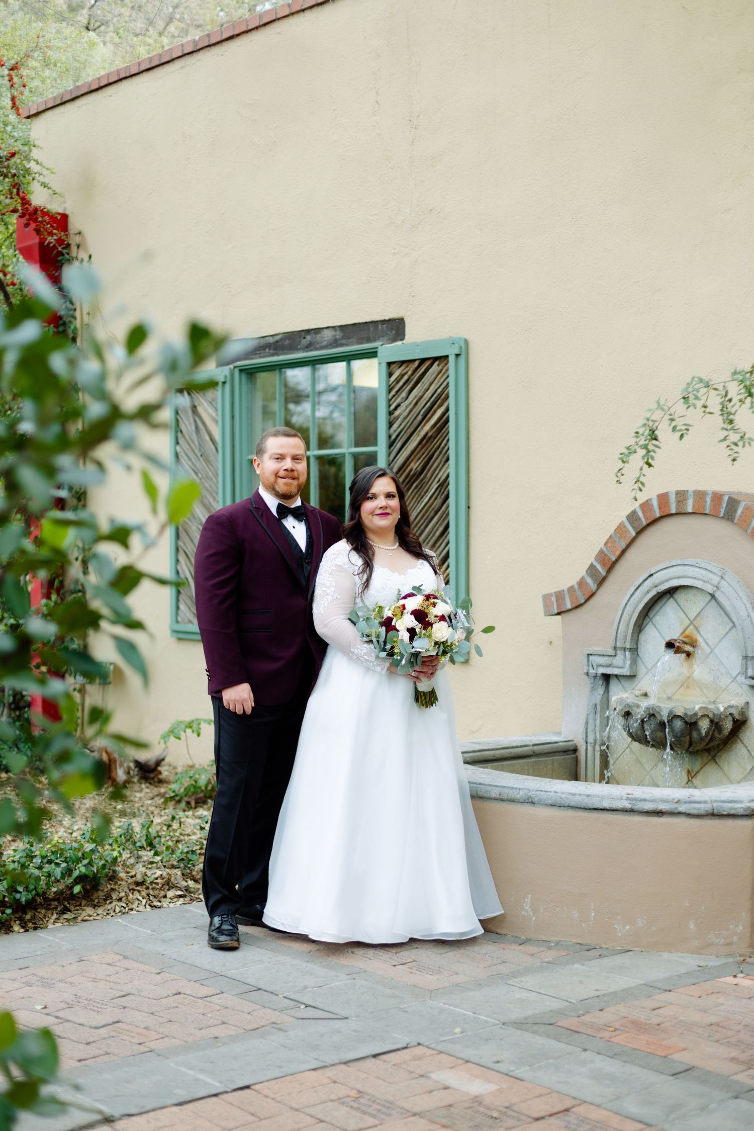 Couple poses for wedding photo against stucco wall with decorative fountain and greenery.