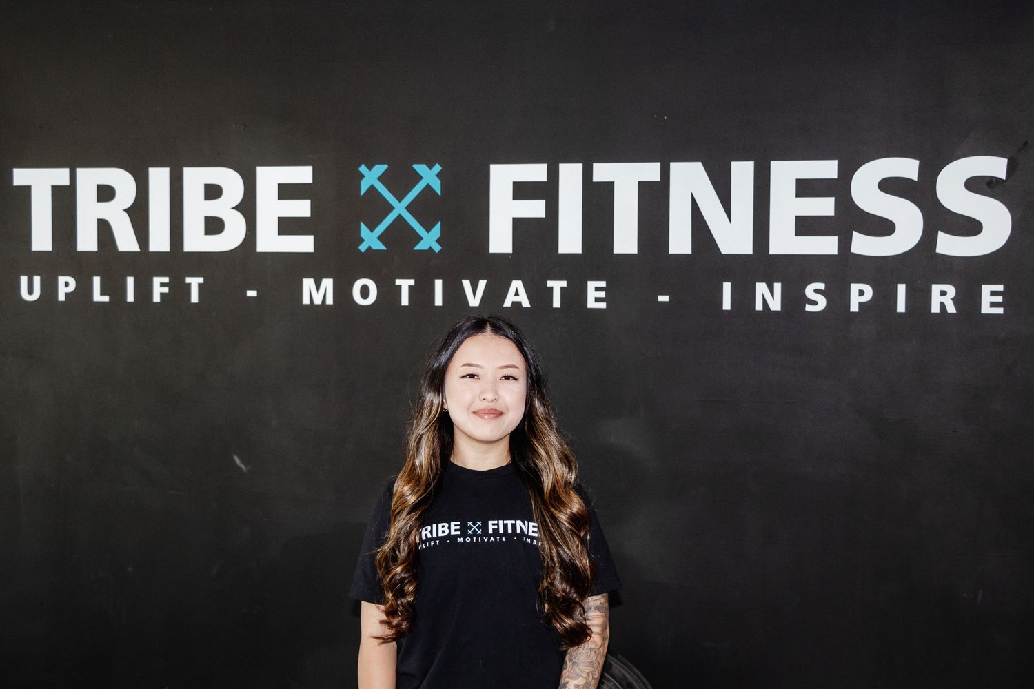 Woman stands in front of a black with with "Tribe Fitness - Uplift, Motivate, Inspire" logo, smiling in a branded t-shirt