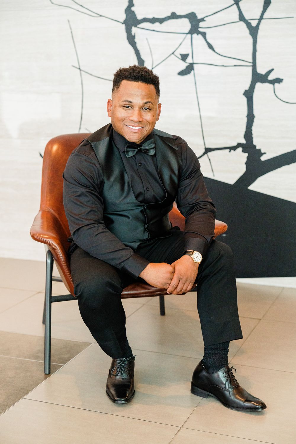 A person wearing a black suit and bow tie sits confidently in a brown leather chair against an artistic wall.