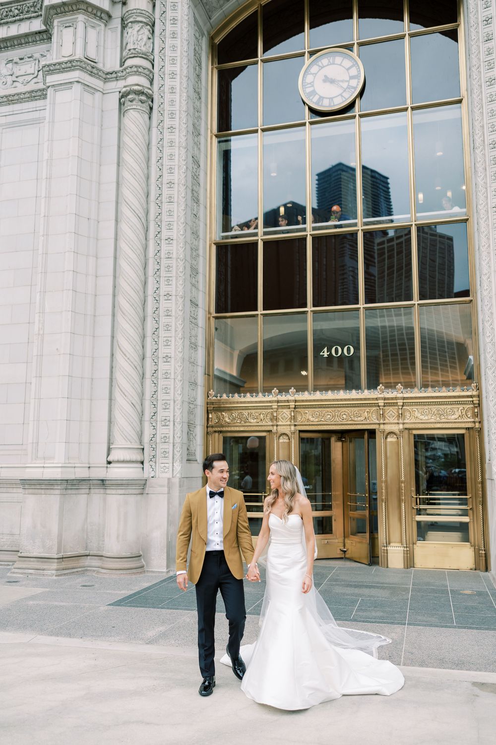 Wedding couple walking in front of grand historic building with glass windows and architectural details.