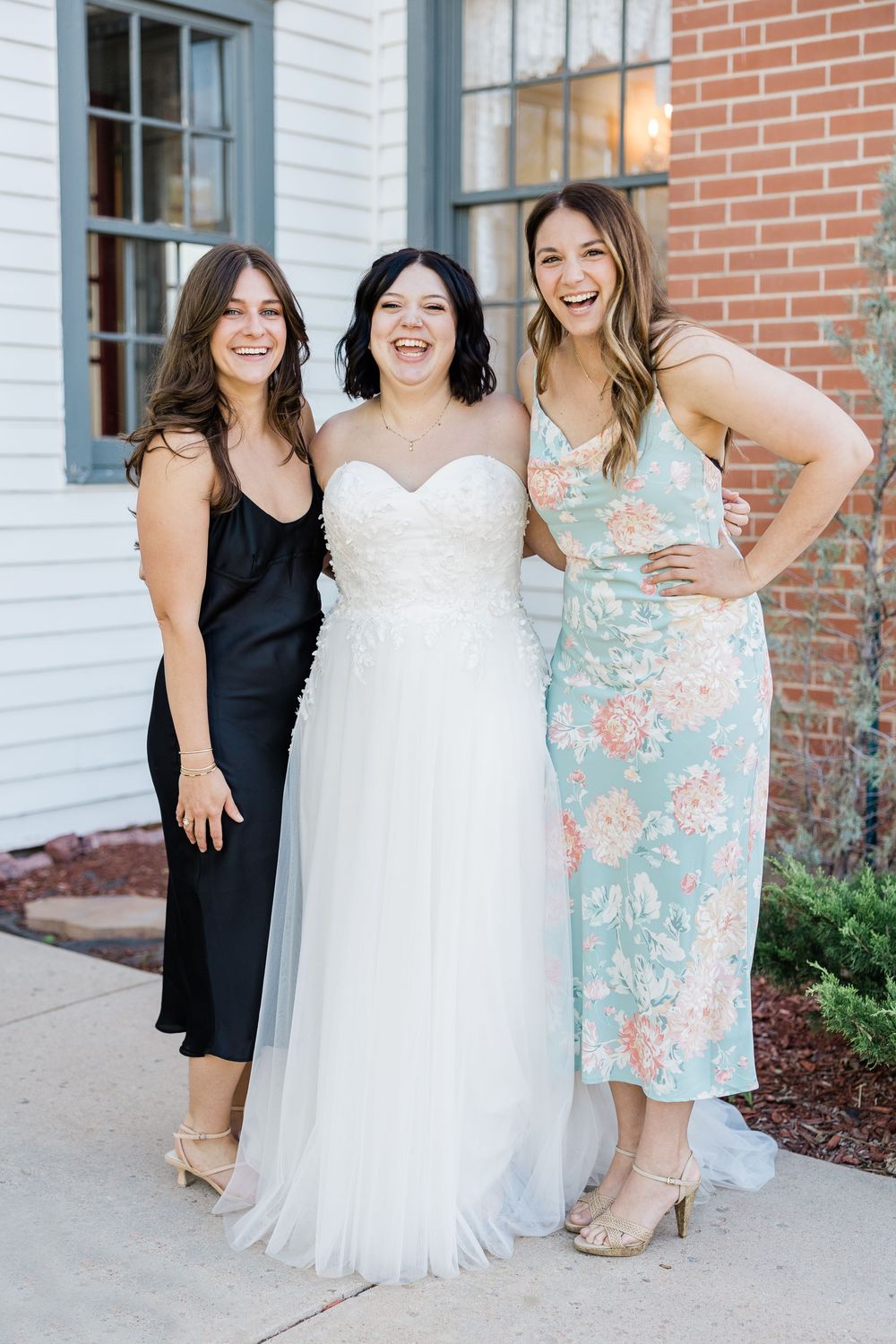 Group photo of bridesmaids in black and floral dresses with bride in white gown outside a brick building.