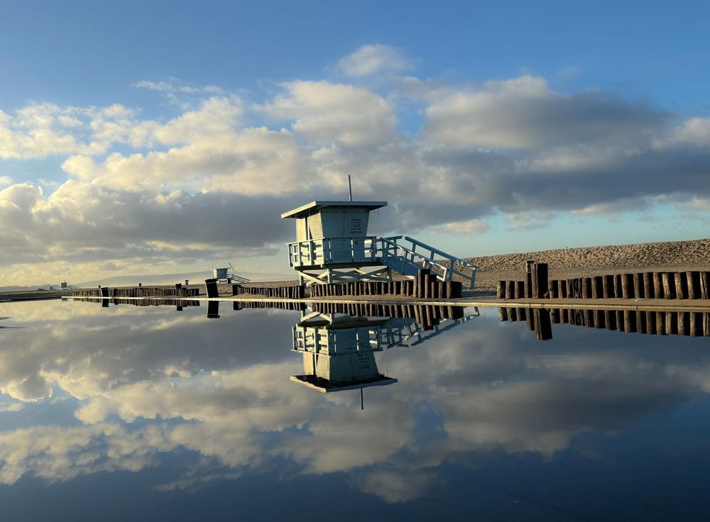 Lifeguard tower reflection in still water with cloudy blue sky and wooden pier structure.
