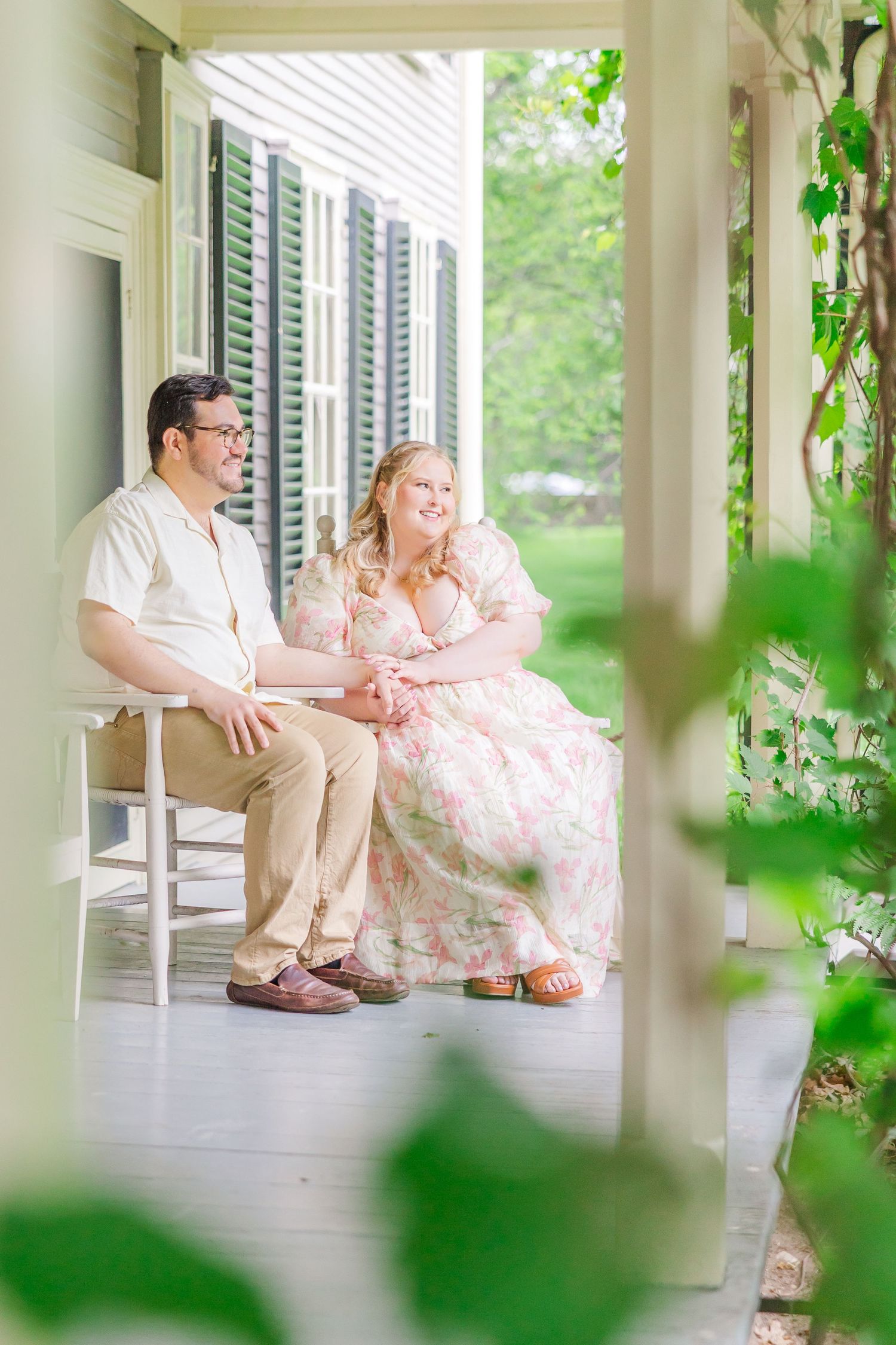 Couple relaxes together on rocking chairs on a sunny white porch with green foliage in the foreground.