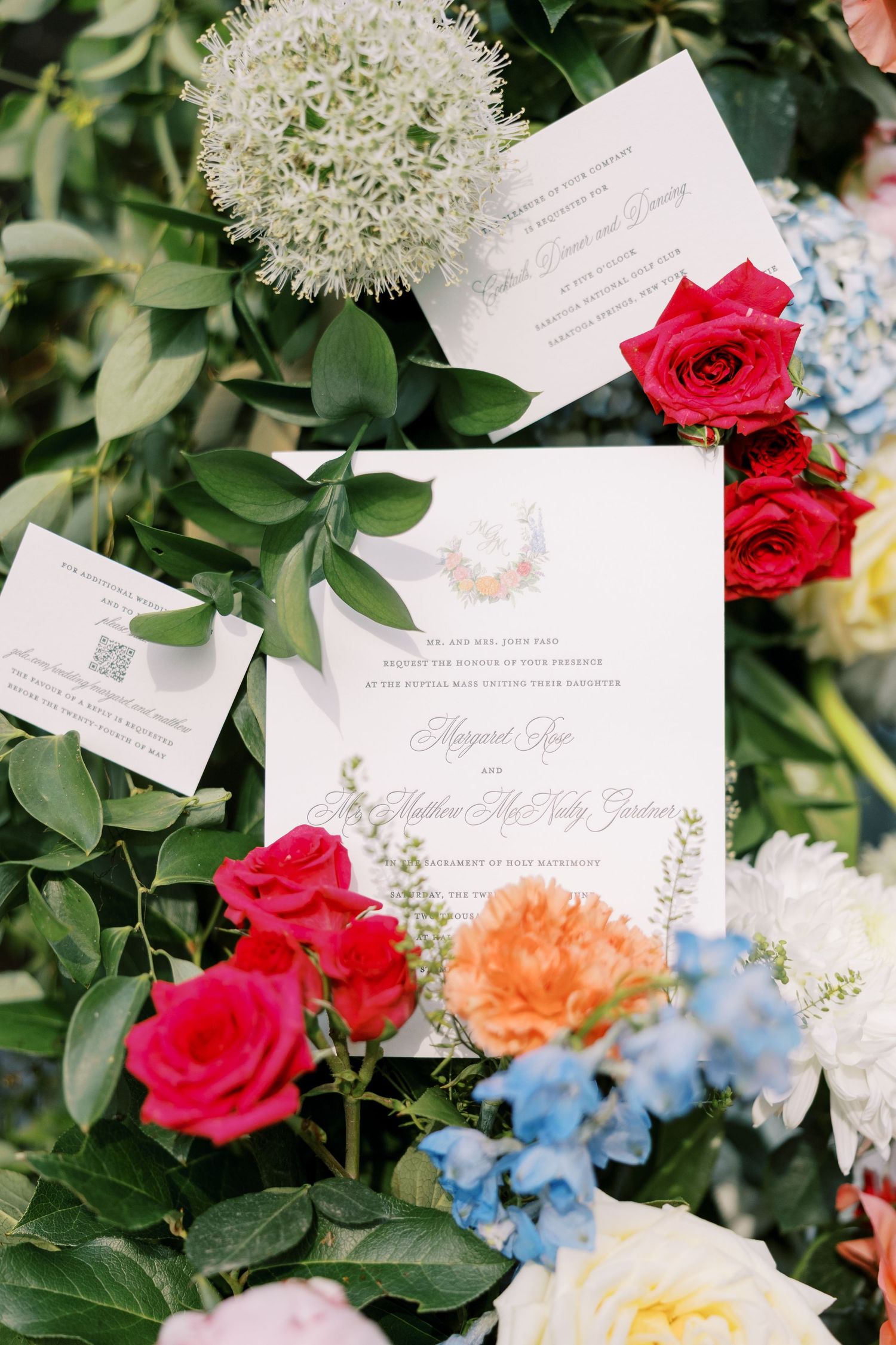 Wedding invitation suite styled with red roses, blue hydrangeas, and coral flowers against lush greenery.