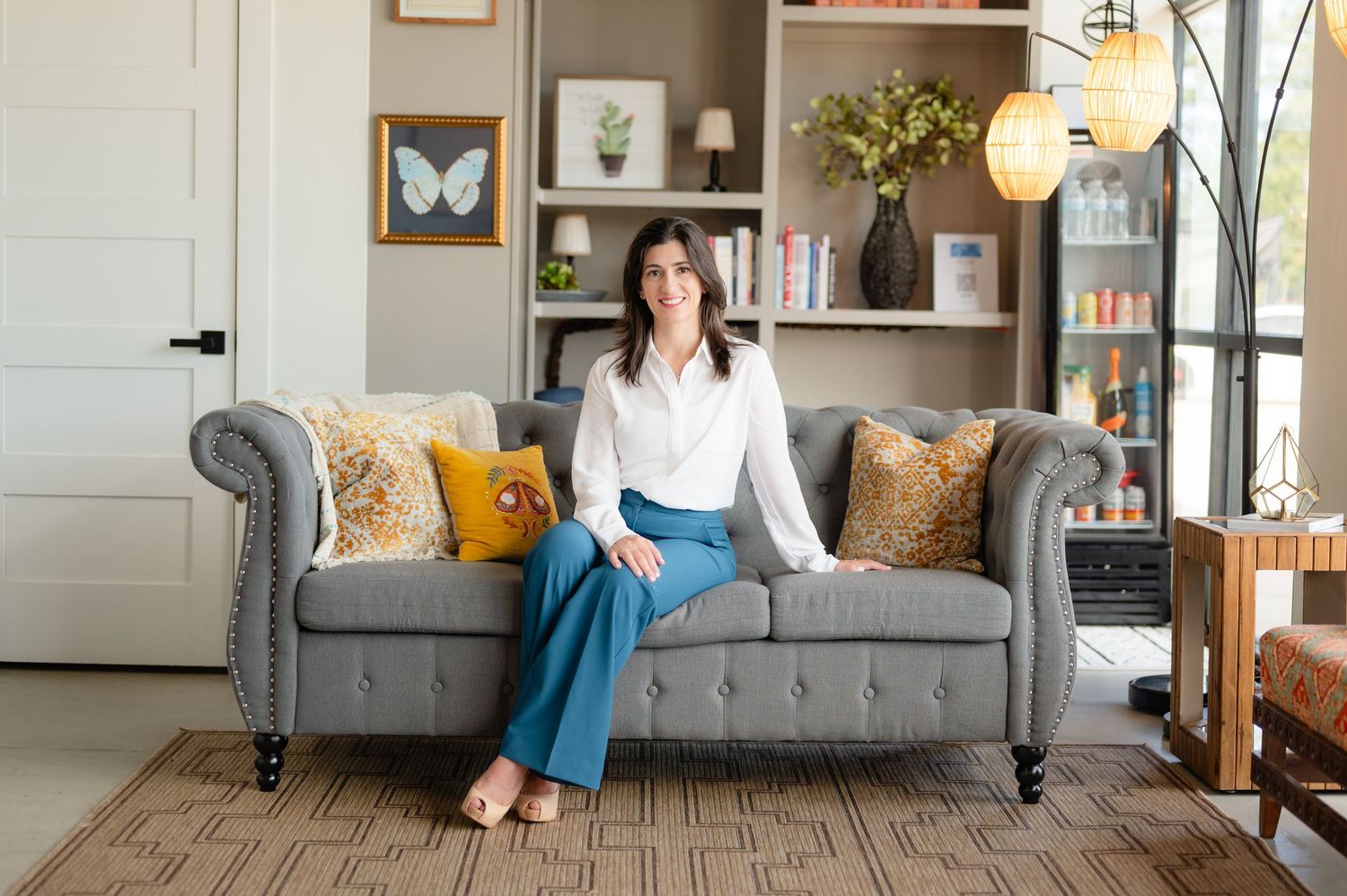 Professional portrait of someone wearing a white button-down shirt and blue pants seated in a yellow accent chair by bookshelves.