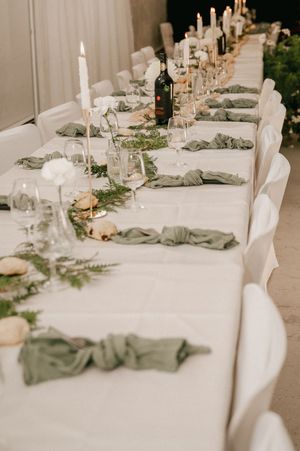 Elegant wedding reception table settings with sage green runners, white linens, and tall taper candles in neutral tones.