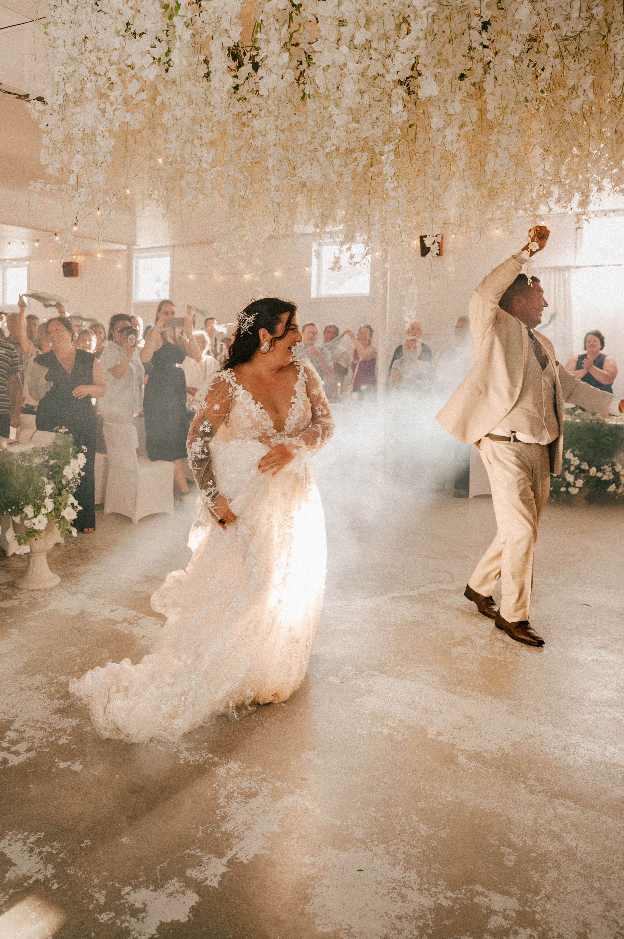 A couple dances their first dance under hanging floral decorations with atmospheric fog effects at an indoor wedding reception.