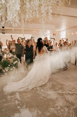 Wedding reception guests gather in an elegant white venue with low-lying fog effect creating a dreamy atmosphere on the dance floor.