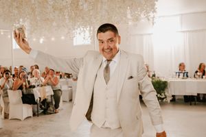A person in a light beige suit and tie celebrates during a wedding reception with guests seated at tables in the background.