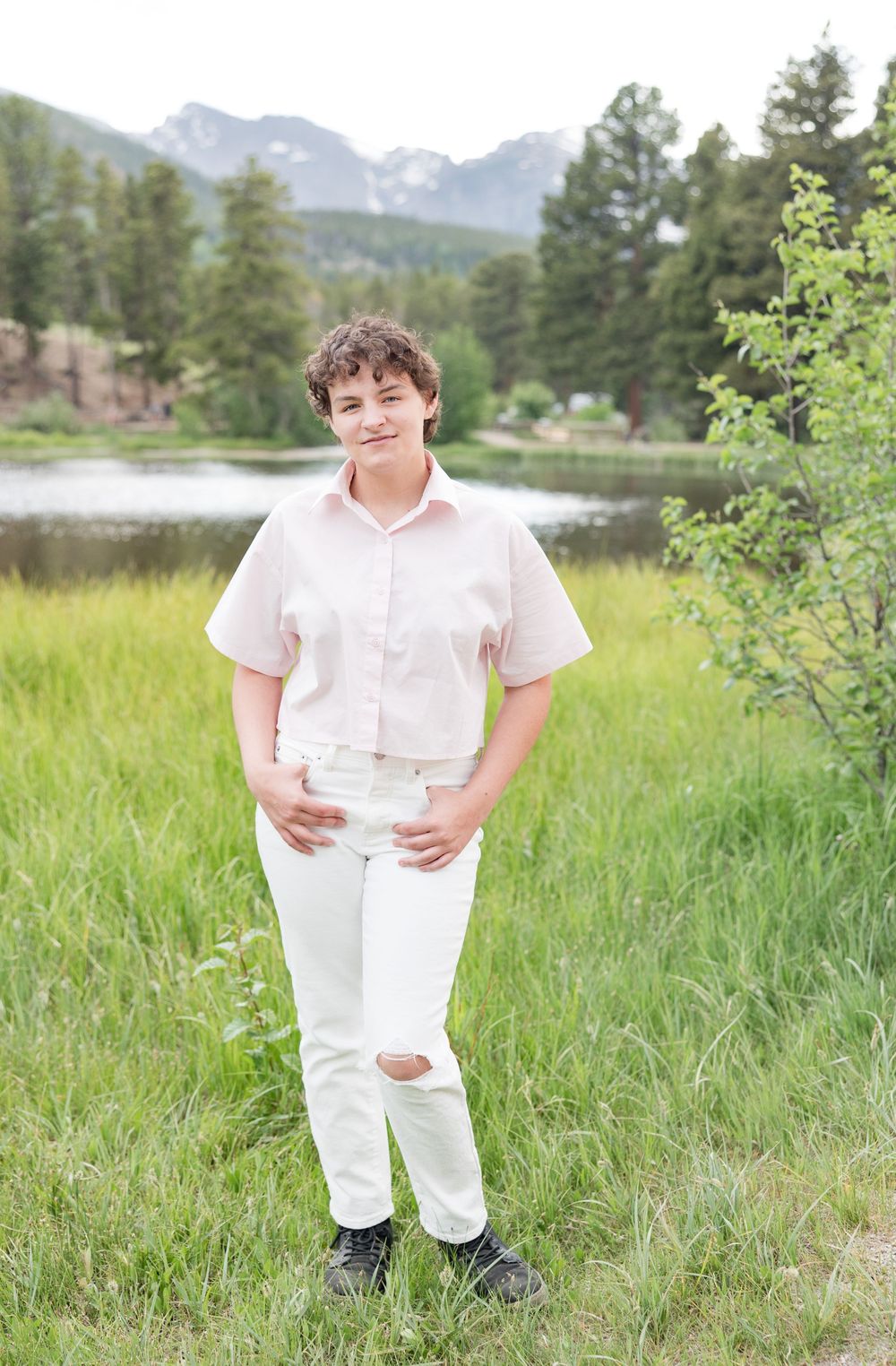 Child in white outfit poses playfully in lush green meadow with scenic mountain backdrop.