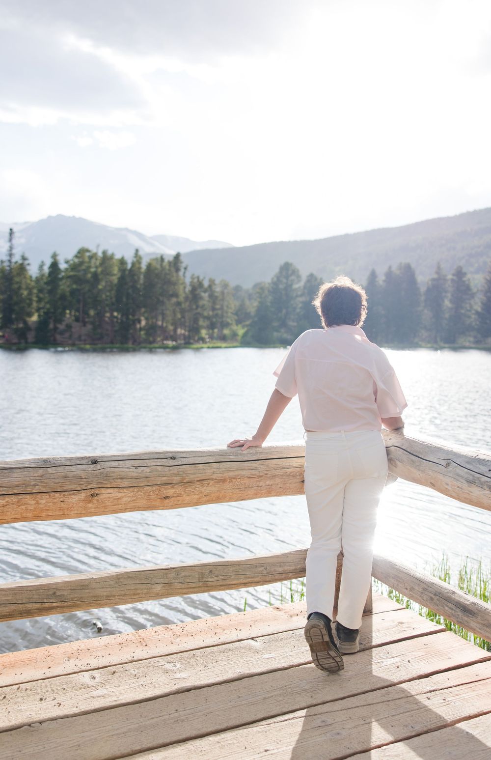 Someone in white clothing leans on a wooden deck railing overlooking a serene mountain lake surrounded by evergreen trees.
