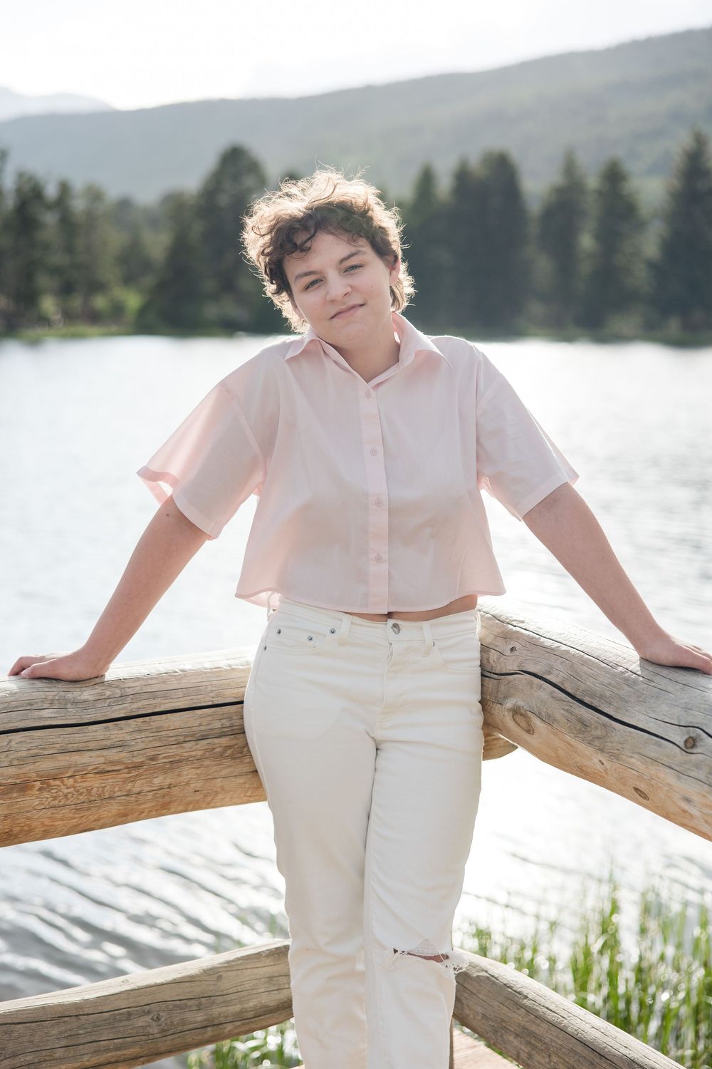 Person wearing white outfit leans on wooden railing by a serene lake with mountains in the background.