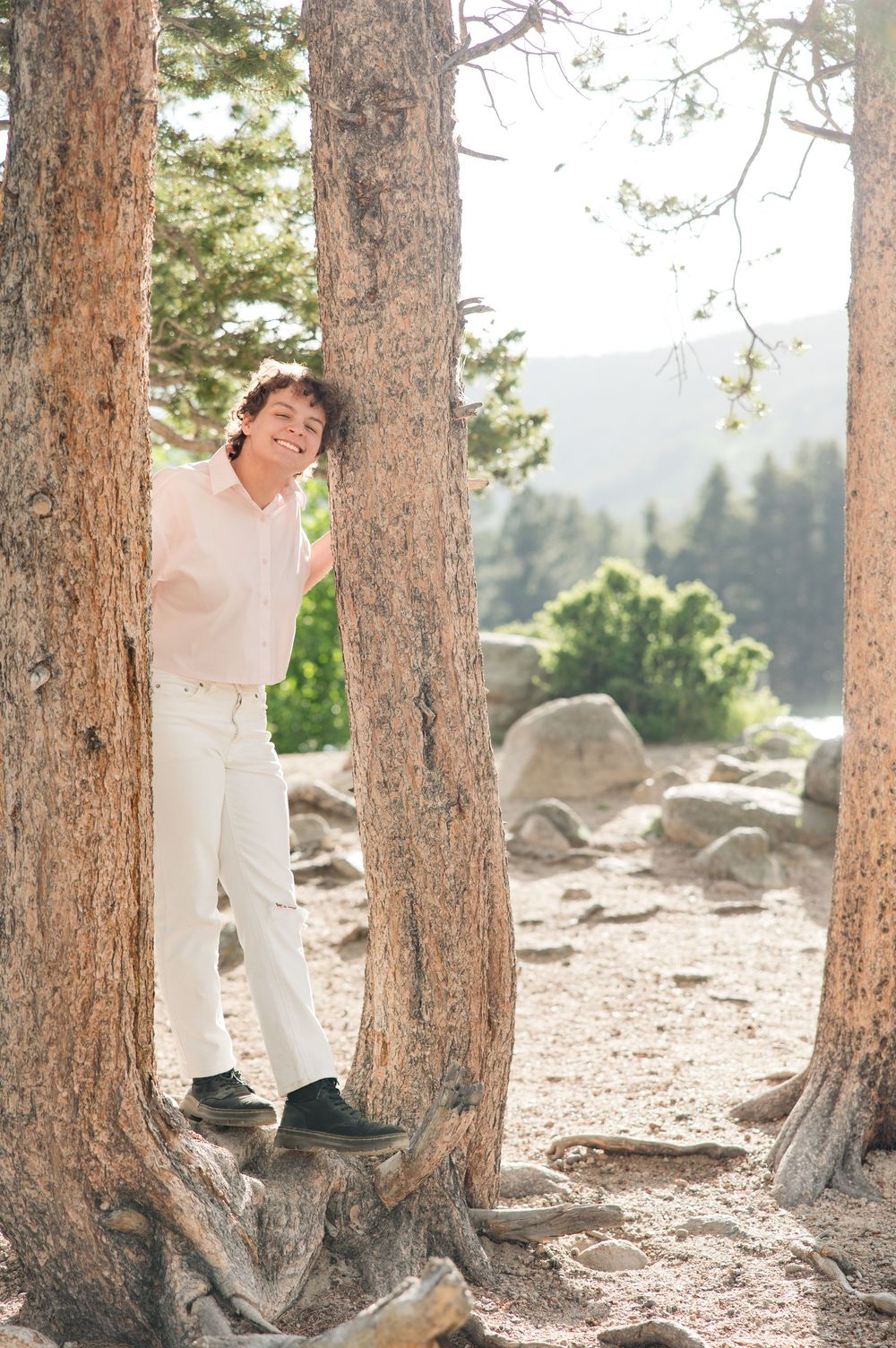 Person in white attire dances joyfully among tall pine trees in a mountainous forest setting.