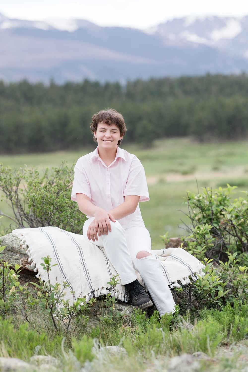 Person in white clothing poses on rocks surrounded by green vegetation and mountain scenery.
