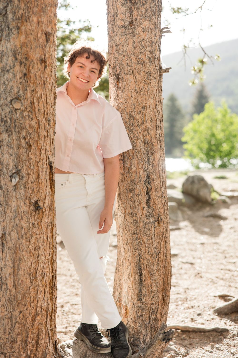 Child in white clothing poses playfully between two tree trunks in a natural outdoor setting during a photo session.