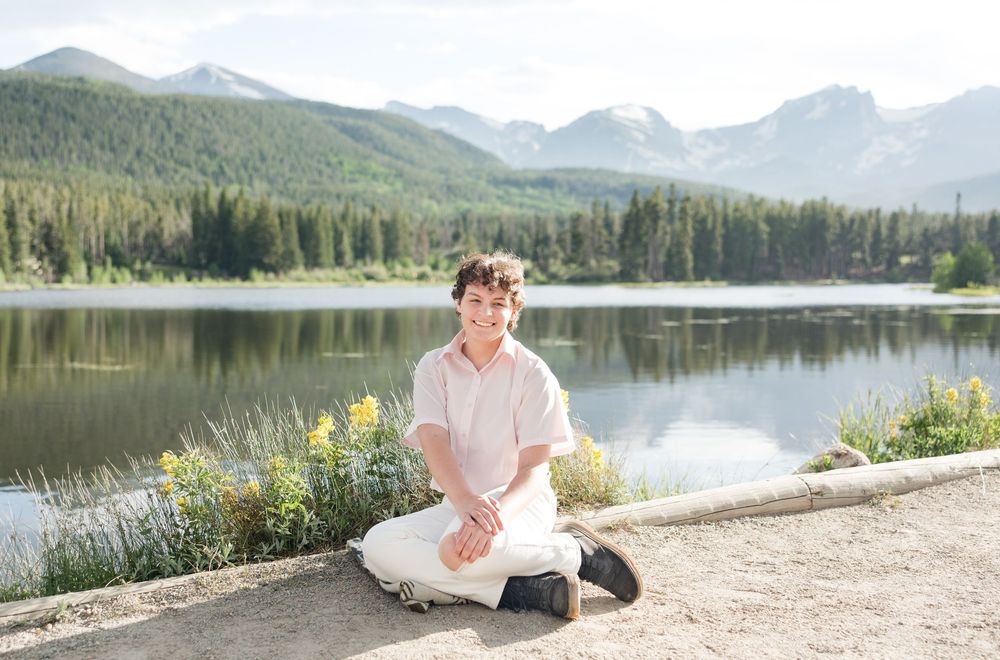 Someone sits by a serene mountain lake surrounded by evergreen forest on a sunny day in the wilderness.
