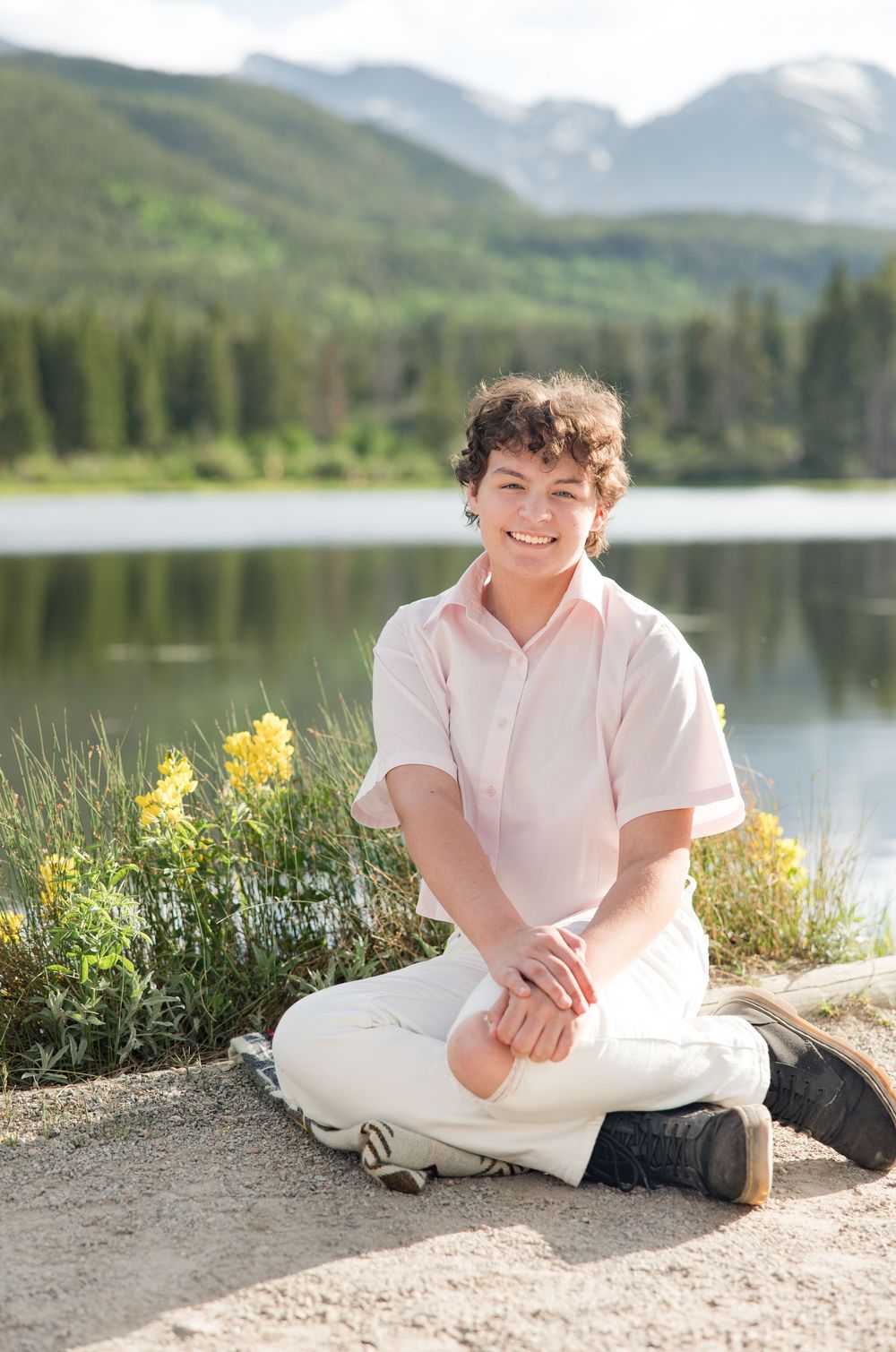 Someone sits cross-legged by a serene mountain lake with wildflowers blooming nearby on a sunny summer day.