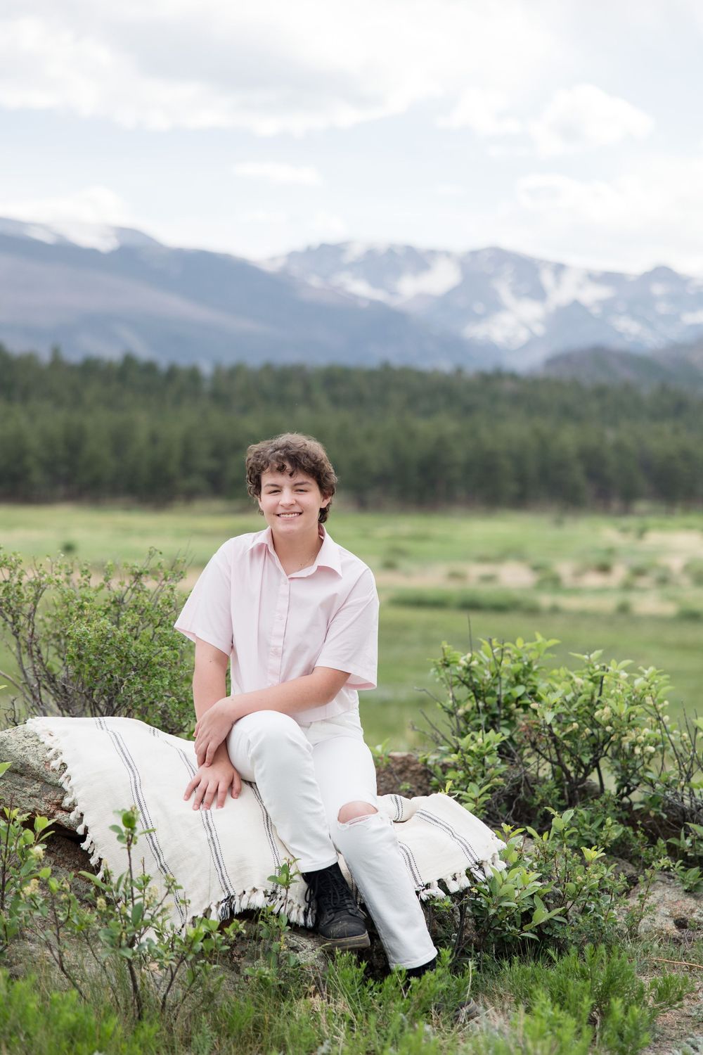 Person in white clothing sitting on rocks with scenic mountain landscape and meadow in background.