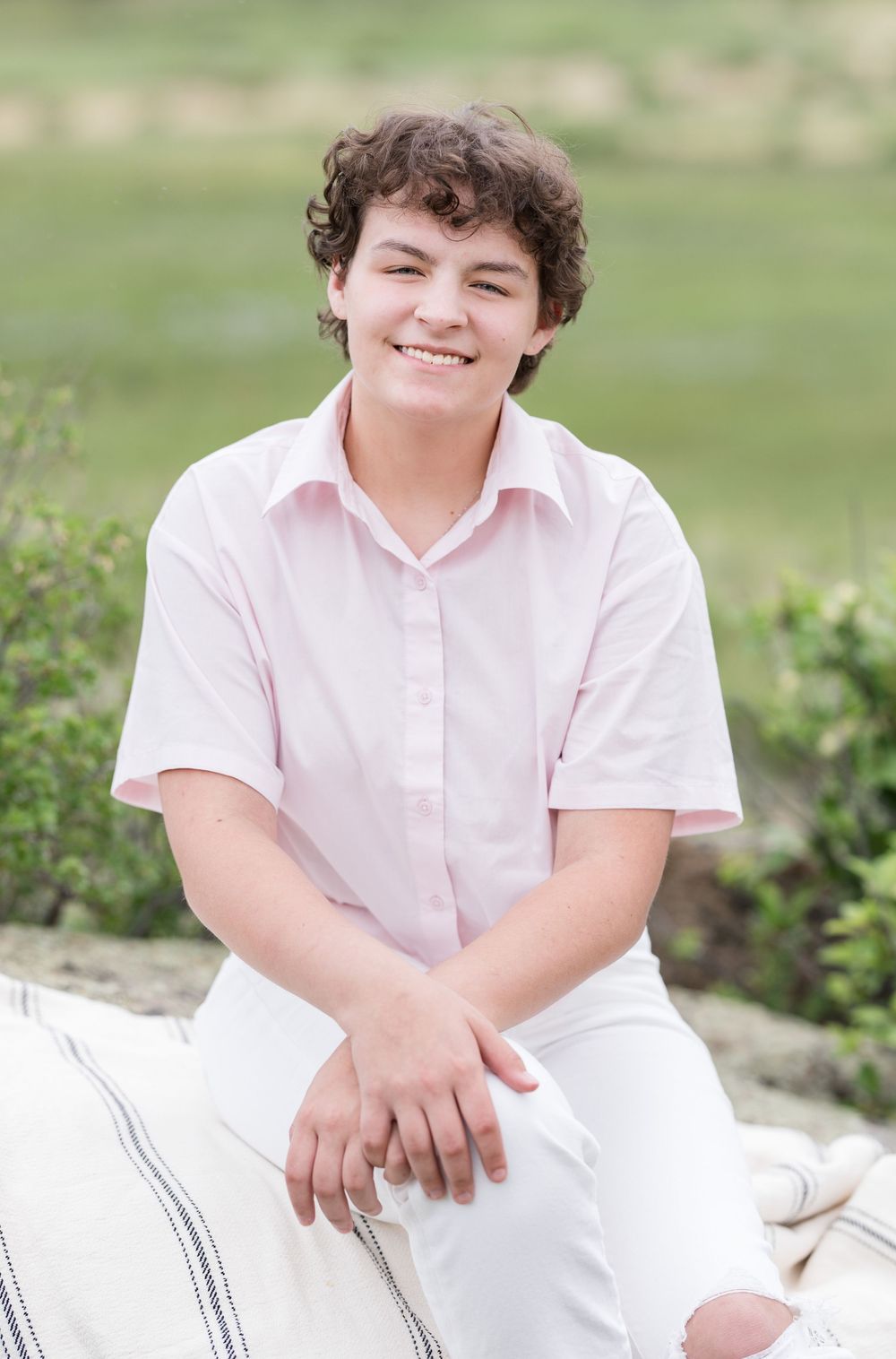 A photo taken outdoors shows someone in a pink short-sleeved shirt and white pants smiling while seated in a garden setting.
