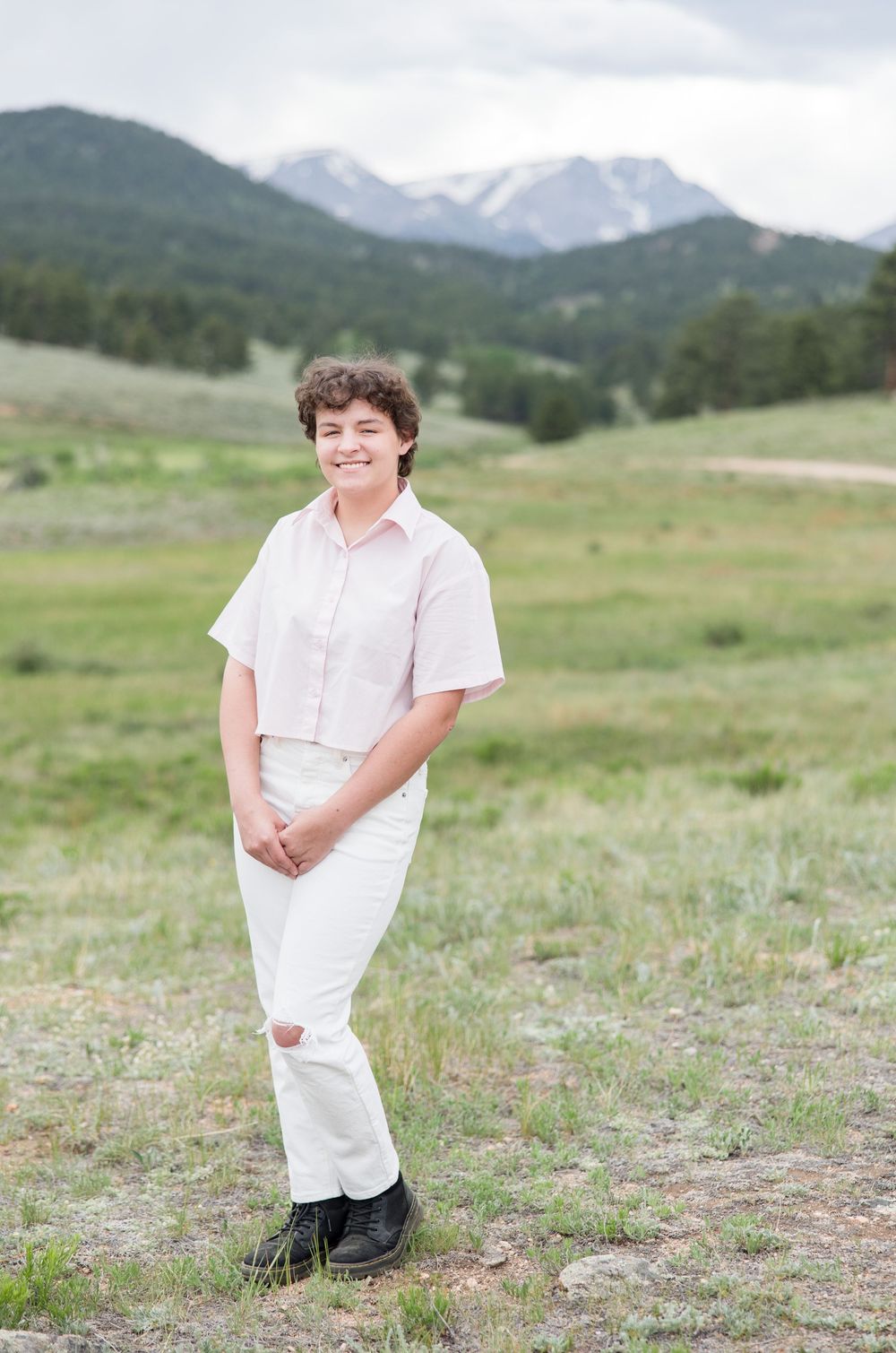 Someone in a white button-down shirt poses in a grassy meadow with snow-capped mountains visible in the background.