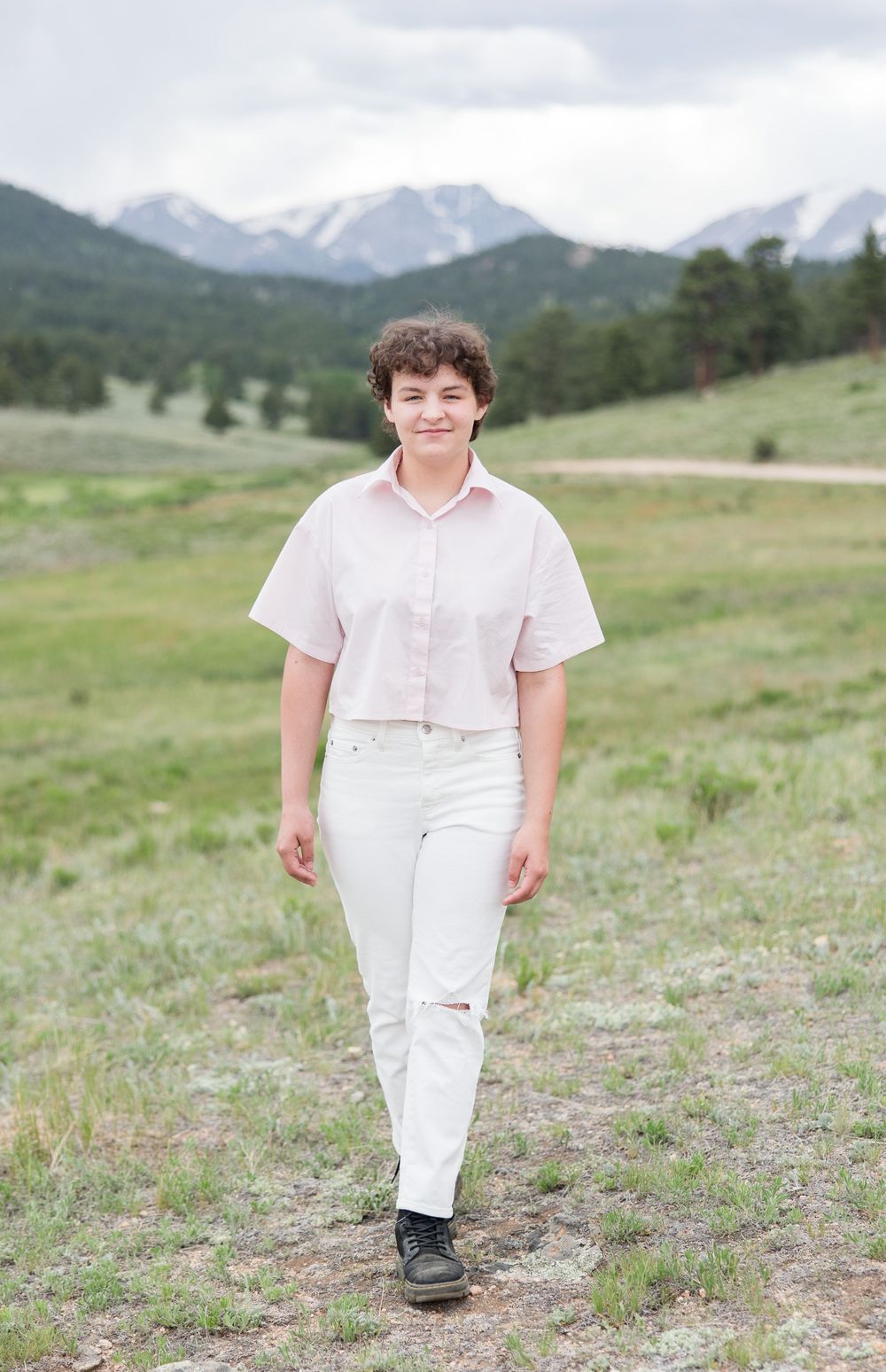 A person in white outfit smiling in a grassy field with scenic mountains in the background.