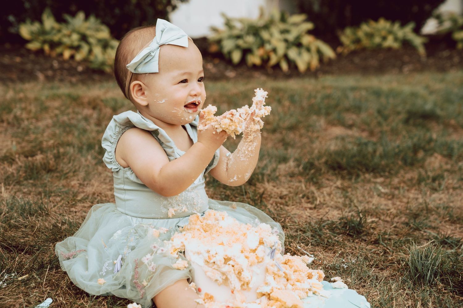 A joyful baby girl in a floral dress and bow headband smashes her first birthday cake outdoors on green grass.