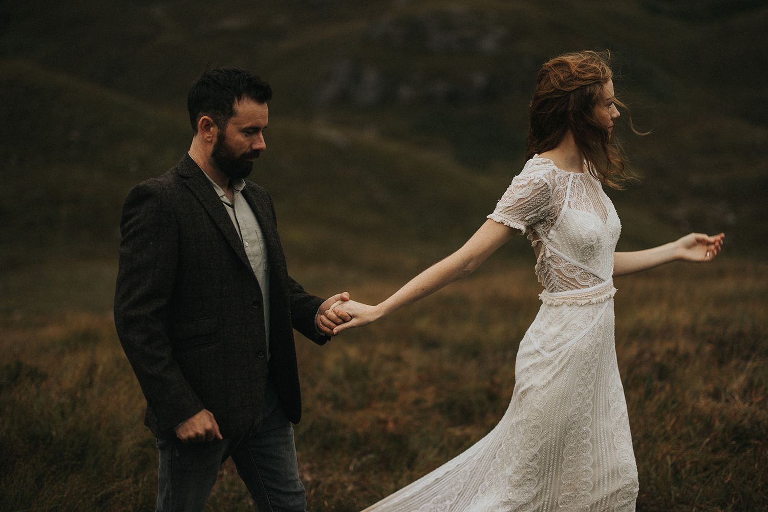 A couple in wedding attire walks together through a misty mountainous field at dusk.