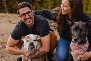 A couple sits outdoors with their two dogs wearing bandanas while sharing a joyful moment together.